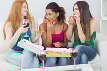 Happy young female friends eating pizza with wine on sofa at home